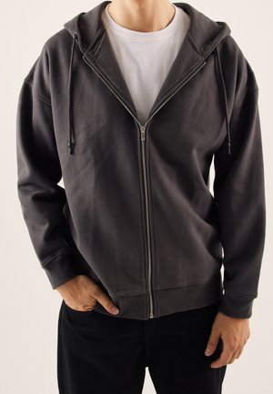 Osoba nosící tmavě šedou mikinu na zip přes bílé tričko, jedna ruka v kapse a druhá volně podél těla.