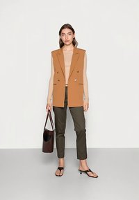 Bruin dubbelgeknopen vest, beige geribd topje, olijfgroene strakke broek, zwarte slip-on sandalen en een bruine schoudertas.