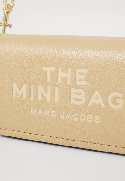 Kreimas ādas mini soma ar embossētu uzrakstu "THE MINI BAG MARC JACOBS." Uzraksts ir balts un ir ievērojami izcelts uz priekšpuses.