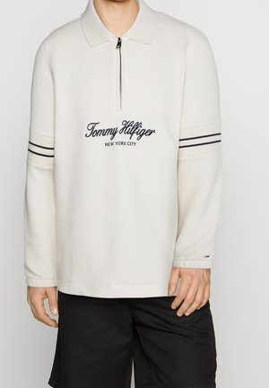 Biała bluza z zamkiem i kołnierzykiem, ozdobiona haftowanym granatowym logo oraz dwoma poziomymi granatowymi paskami na rękawach. Gładka tekstura.