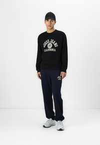 Sudadera negra con el logo "Guess Jeans California"; combinada con pantalones de chándal azul marino y zapatillas grises; el modelo lleva una gorra negra.