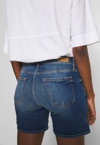 Pantalones cortos de mezclilla en azul oscuro con un diseño ajustado, que cuentan con dos bolsillos traseros y una etiqueta de cuero en la cintura. Parte de una blusa blanca es visible.