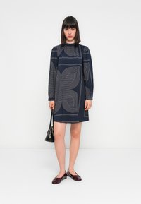 Donna in piedi contro uno sfondo bianco, indossa un abito blu navy con motivi geometrici bianchi, tiene una borsa nera e delle scarpe basse bordeaux.