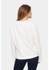 Felpa leggera color beige con maniche lunghe e scollatura rotonda. Presenta un orlo e polsini a coste. Indossata sopra jeans in denim scuri.