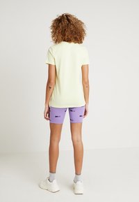 Gul bomullströja i kombination med lila Nike-shorts med ett upprepat logotypmönster. Vita sneakers och ankelsockar fulländar outfiten.