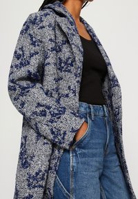 Vrouw in een blauw-wit gebreid jas met patroon, zwarte top en hoge taille blauwe spijkerbroek, met één hand in haar zak.