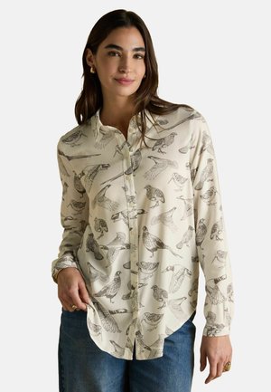 Woman wearing cream button-up shirt with detailed black bird illustrations, paired with blue jeans and gold hoop earrings.