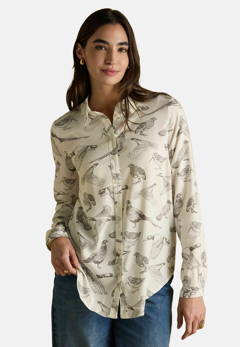 Woman wearing cream button-up shirt with detailed black bird illustrations, paired with blue jeans and gold hoop earrings.