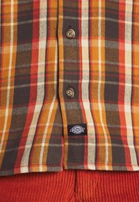 Brązowa, pomarańczowa i kremowa koszula w kratę zapinana na guziki, z dwoma widocznymi guzikami i czarną metką z logo Dickies nad rdzawej barwy spodniami sztruksowymi.