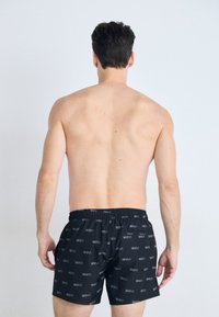 Svart svømmeshorts med et repetitivt logo-mønster, elastisk midjebånd og en avslappet passform. Stoffet ser glatt og lett ut.