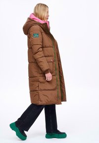 Brązowa puchowa kurtka z kapturem, różowym podszyciem, zielonym zamkiem błyskawicznym i bocznymi kieszeniami; noszona na czarnych spodniach i zielonym obuwiu.