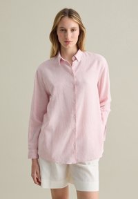 Pink, long-sleeve button-up shirt made of textured fabric, featuring a pointed collar and straight-cut hem, paired with white shorts.