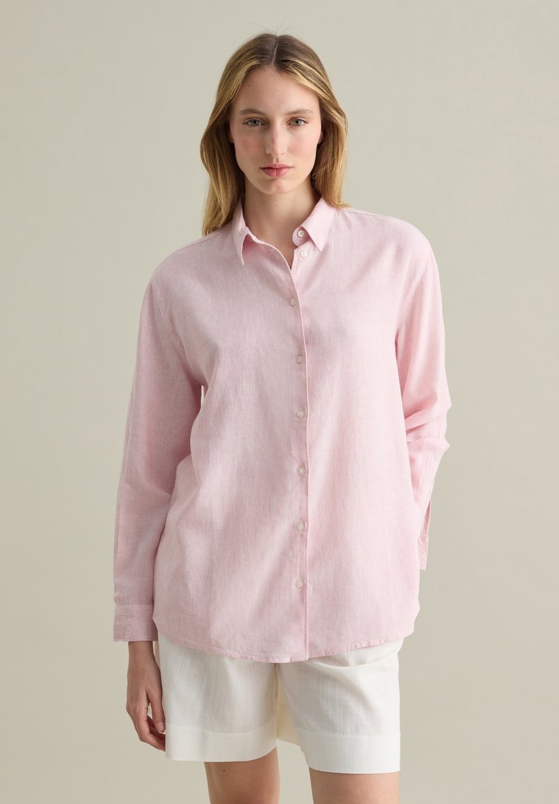 Pink, long-sleeve button-up shirt made of textured fabric, featuring a pointed collar and straight-cut hem, paired with white shorts.