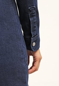 Tummansininen farkkutakki lähikuvassa, jossa näkyy nappi hihansuussa, ompeleet ja teksturoitu pinta. Kangas näyttää hieman kuluneelta.