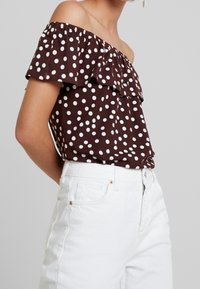 Ruskea pilkullinen olkapään yli menevä paita, jossa on röyhelökaulus, yhdistetty valkoisiin korkeavyötäröisiin farkkuihin. Pehmeä kangas, rento malli, lyhyet hihnat.