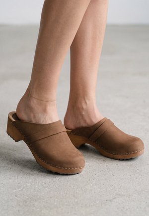 Ben iført brune ruskindstræskostøvler med træsåler på en grå betonbelægning, den ene fod pyntet med en tynd guldfodkæde.