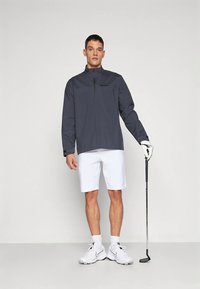 Golf shirt in dark gray, featuring a half zipper and long sleeves, paired with white shorts, white athletic shoes, and holding a golf club.