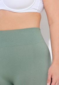 Jasnozielone, prążkowane legginsy z wysokim stanem, ukazujące teksturowany materiał i gładkie wykończenie, uzupełnione białym biustonoszem sportowym powyżej.