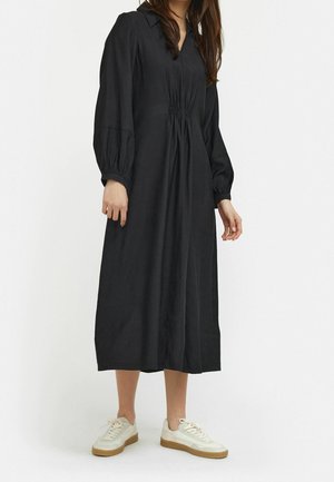 Black long-sleeve dress with a v-neck, gathered waist, and wrist cuffs, made from soft fabric; paired with white sneakers.