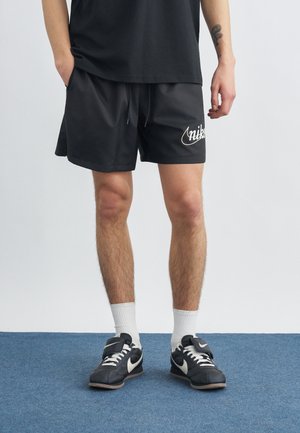 Person wearing black Nike shorts, black and white Nike sneakers with white laces, white socks, and a black shirt standing on blue carpet.