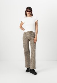 Top bianco a maniche corte abbinato a jeans color marrone chiaro con stampa leopardata e stivali neri chunky, modello in piedi contro uno sfondo bianco semplice.