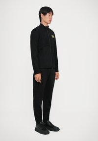 Giovane uomo in piedi, indossa una giacca nera con zip con un piccolo logo sul petto, pantaloni neri e sneaker nere su uno sfondo neutro.