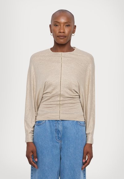 Beige langermet topp med rund hals, samlet tekstur i midjen, og en sentral søm. Kombinert med løse blå denimbukser.