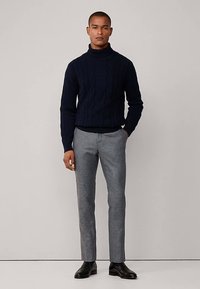 Námořnicky modrý rolák s kabelovým vzorem spojený s šedými kalhotami na míru a černými koženými botami, na neutrálním pozadí.