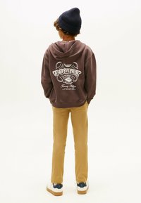 Sudadera marrón con el logo blanco "TOMMY" en la parte posterior, combinada con pantalones beiges y zapatillas blancas con detalles en azul marino. Gorro de punto en la cabeza.