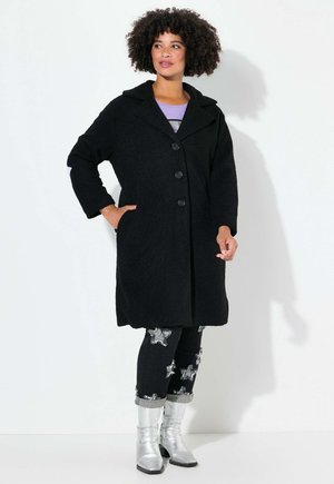 Black, textured wool coat with a notched collar and three buttons; worn over dark jeans with silver star embellishments and metallic boots.