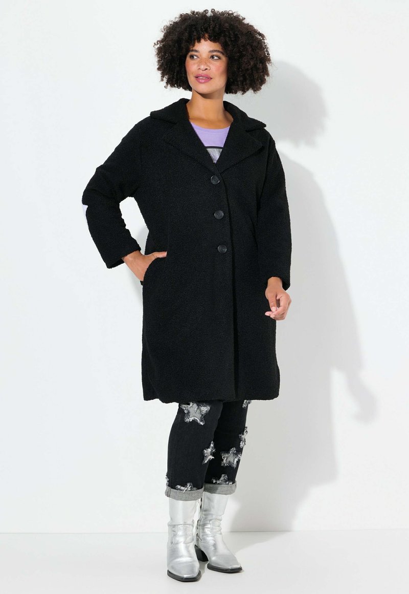 Cappotto di lana nero e textured con colletto a scialle e tre bottoni; indossato sopra jeans scuri con decorazioni a forma di stella argentata e stivali metallici.