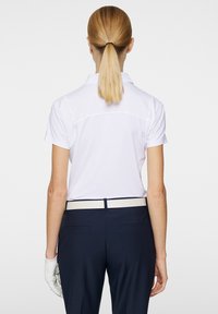 Valkoinen lyhythihainen polo-paita, jossa on teksturoitu pinta, yhdistettynä tummansinihin housuihin ja eriväriseen vaaleaan vyöhön. Valkoinen golf-käsine on näkyvissä.