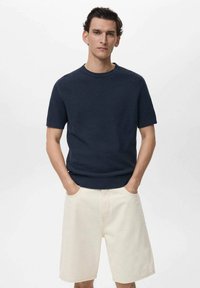 Maglione navy a maniche corte in maglia con un design testurizzato, abbinato a pantaloni corti in denim beige chiaro. La modella ha le mani in tasca e si trova di fronte a uno sfondo bianco.
