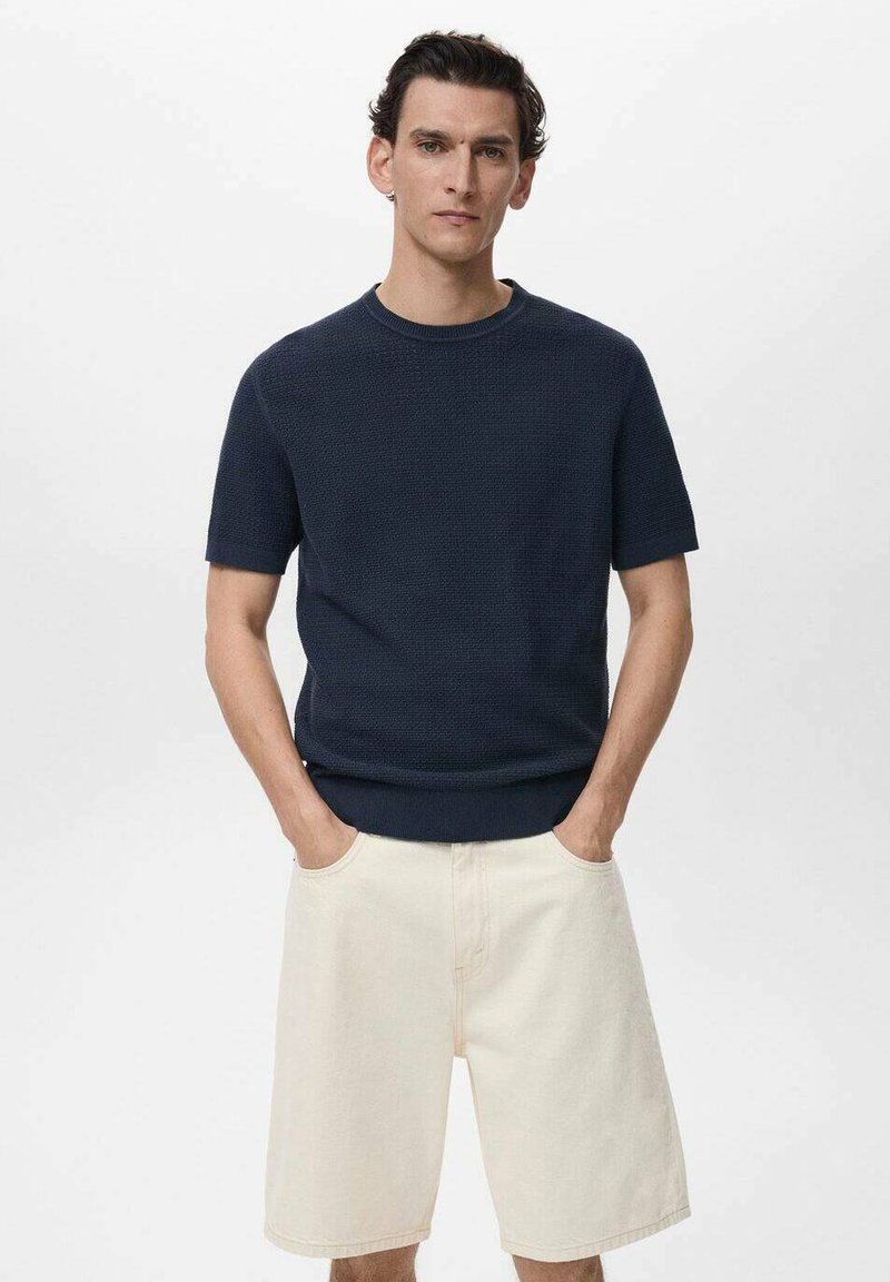Maglione navy a maniche corte in maglia con un design testurizzato, abbinato a pantaloni corti in denim beige chiaro. La modella ha le mani in tasca e si trova di fronte a uno sfondo bianco.