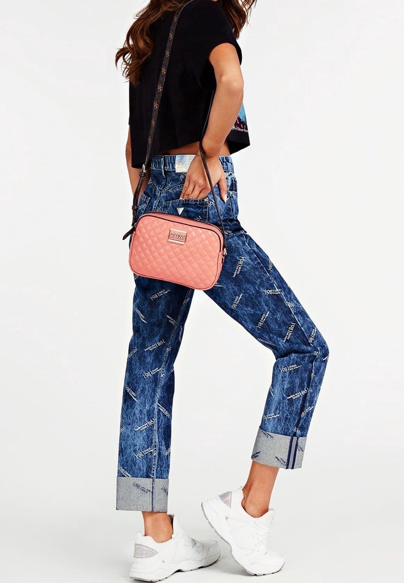 Světlá růžová prošívaná kabelka přes rameno s logem značky, doplněná modrými džínovými kalhotami s potiskem a zkráceným lemem. Bílé tenisky.