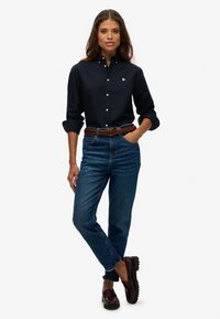 Camisa de botón azul marino, jeans de tiro alto en color azul, cinturón trenzado marrón y mocasines oscuros; silueta elegante y líneas limpias.