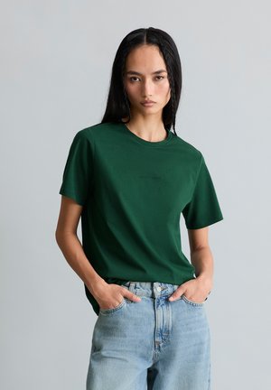 Junge Frau mit langen dunklen Haaren, die ein dunkelgrünes kurzärmeliges T-Shirt und hellblaue Jeans trägt, steht mit den Händen in den Taschen.
