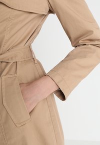 Trench-coat beige avec une taille ceinturée, doté d'un grand col et d'une poche latérale unique, fabriqué en tissu lisse et durable avec un subtil éclat.