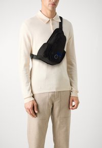 Svart skulderveske i nylon, med en glidelåslomme foran, blå etikettdetalj, og justerbar stropp, båret over brystet.