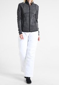Woman walking wearing a grey zip-up jacket, white insulated snow pants, and black boots against a plain white background.