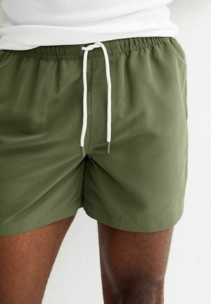 Uomo che indossa pantaloncini verde oliva con coulisse bianca e maglietta bianca a costine, cosce visibili contro uno sfondo chiaro semplice.