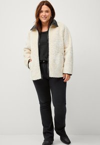 Crèmekleurige, met fleece gevoerde jas met een zwarte kraag. Beschikt over een voorkant met rits, twee zijzakken en geribbelde details op de manchetten.