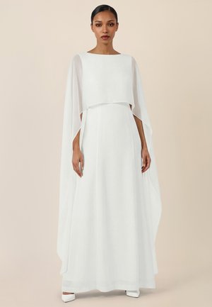 Frau mit zurückgebundenen Haaren, die ein langes weißes Kleid mit fließenden Umhangärmeln trägt, steht vor einem schlichten beigen Hintergrund.