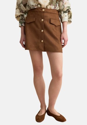 Minigonna in suede marrone con bottoni frontali e tasche con patta, indossata con una camicetta floreale e ballerine in suede marrone abbinate.