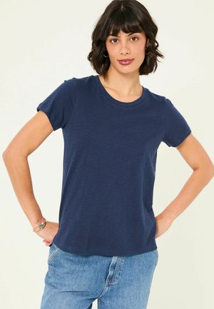 Woman with short dark hair wearing gold hoop earrings, navy blue t-shirt, and light blue jeans, posing with hands on hips against a plain background.