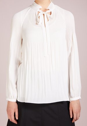 Vrouw draagt een witte blouse met lange mouwen, plooien en een gebonden strik bij de hals, gecombineerd met een zwarte rok.