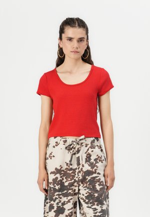 Haut rouge à manches courtes avec un décolleté arrondi, fabriqué à partir d'un tissu doux. Associé à un pantalon beige présentant un motif de taches brunes irrégulières.