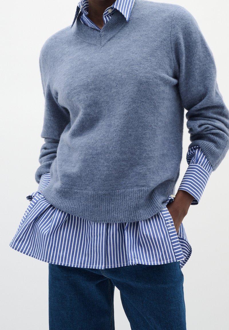 Personne portant un pull bleu col en V par-dessus une chemise à rayures bleu et blanc, avec un jean bleu foncé, les mains dans les poches.