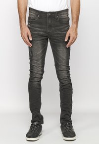 Man wearing slim-fit black distressed jeans and black sneakers standing against a plain light background.