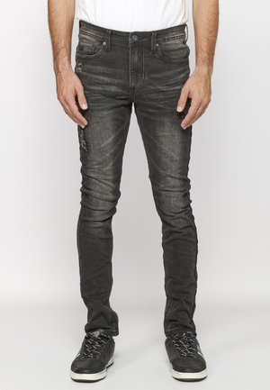 Hombre vestido con jeans ajustados negros desgastados y zapatillas negras, de pie contra un fondo claro y simple.